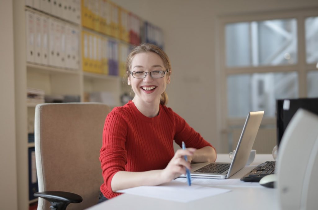 Uśmiechnięta kobieta pracuje przy biurku. Ma przed sobą laptop, a w dłoni trzyma długopis. 