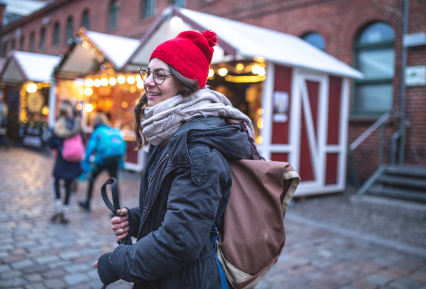 Zima. Dziewczyna w czerwonej czapce idzie wzdłuż handlowych, świątecznie przystrojonych budek. Odwraca się do tyłu, w kierunku osoby robiącej zdjęcie.