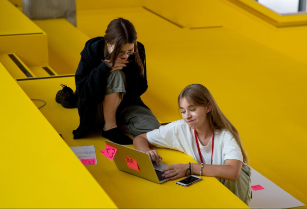 Dwie nastolatki pracują przy laptopie. Siedzą wygodnie w żółtym, nowoczesnym holu.